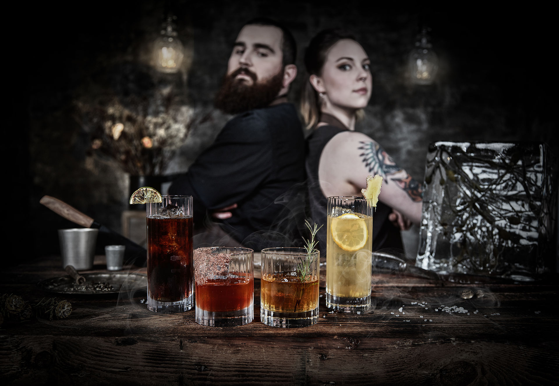 Vier Gläser mit unterschiedlichen Drinks stehen auf einer Holzplatte, im dunklen Hintergrund sind ein Mann und eine Frau zu sehen.